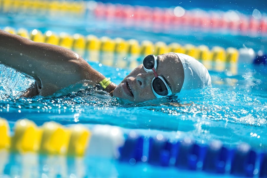 Close-up photo of female swimmer in the motion in the swimming pool outdoors. She wears a black-lime swimsuit, a white swim cap and swim glasses. Girl looks into the camera. Horizontal.