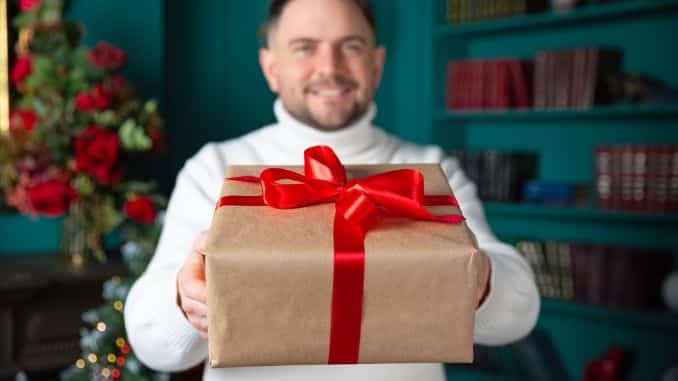 Happy young man at home holding Christmas gift. Happy young man in white sweater at home holding Christmas gift